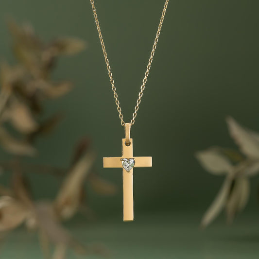 A gold cross pendant with a heart-shaped gemstone is suspended from a chain against a blurred green background.
