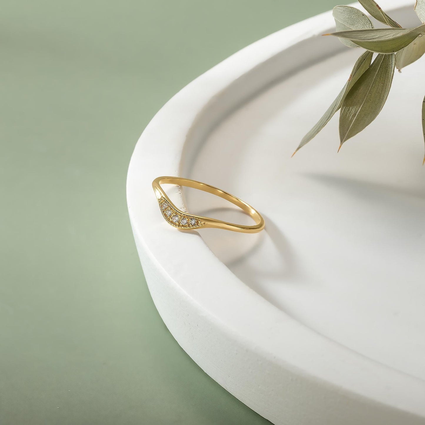 A gold ring with a diamond-like stone is placed on a white, curved surface, with a plant leaf visible in the background.