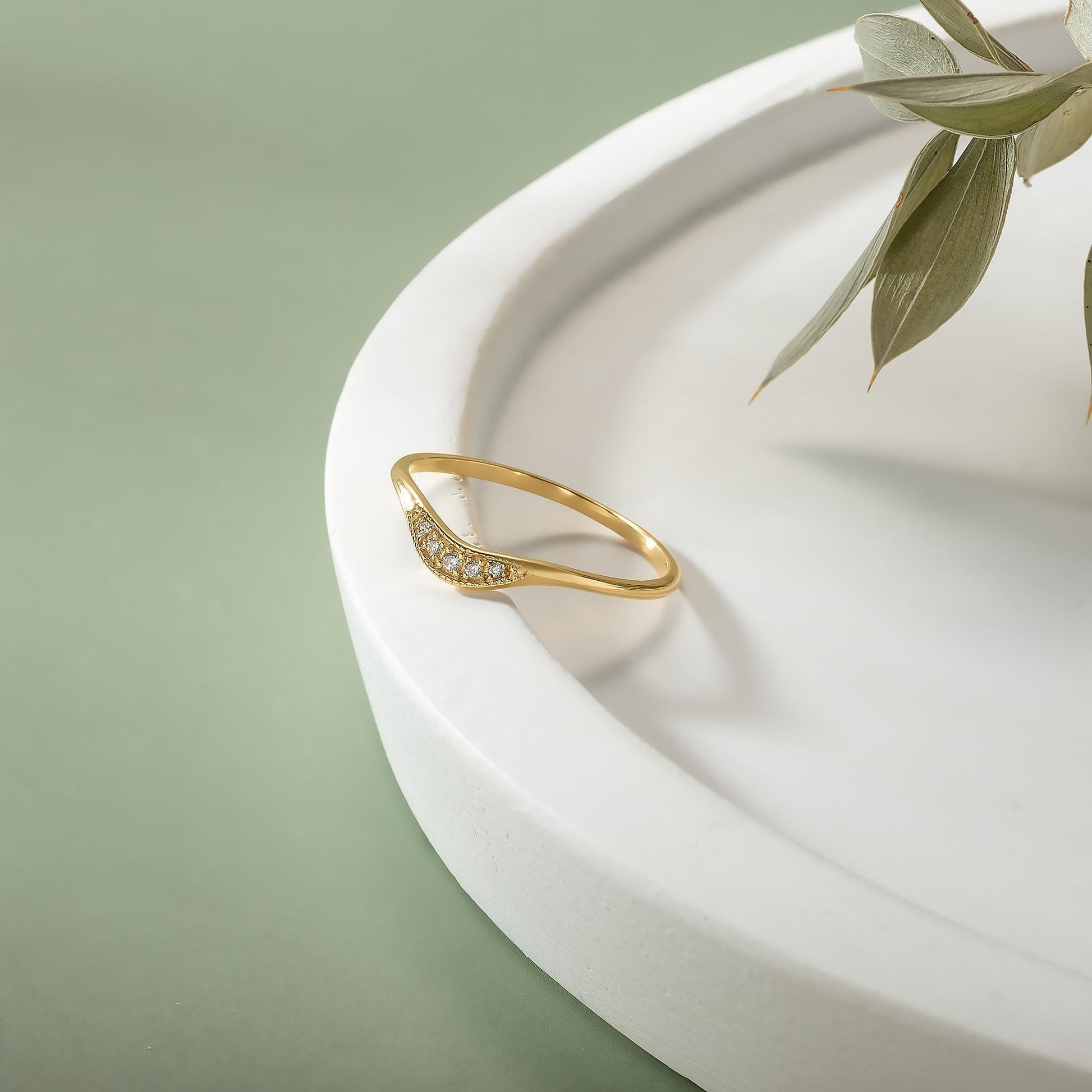 A gold ring with a diamond-like stone is placed on a white, curved surface, with a plant leaf visible in the background.