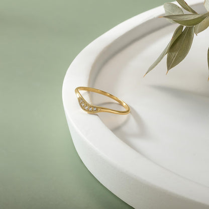 A gold ring with a diamond-like stone is placed on a white, curved surface, with a plant leaf visible in the background.