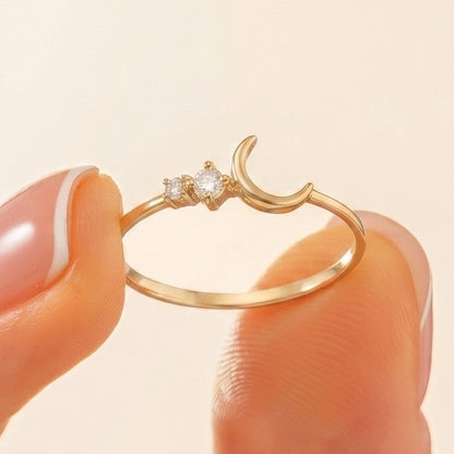 A gold ring with a crescent moon and two small diamonds is being held between a person's fingers.