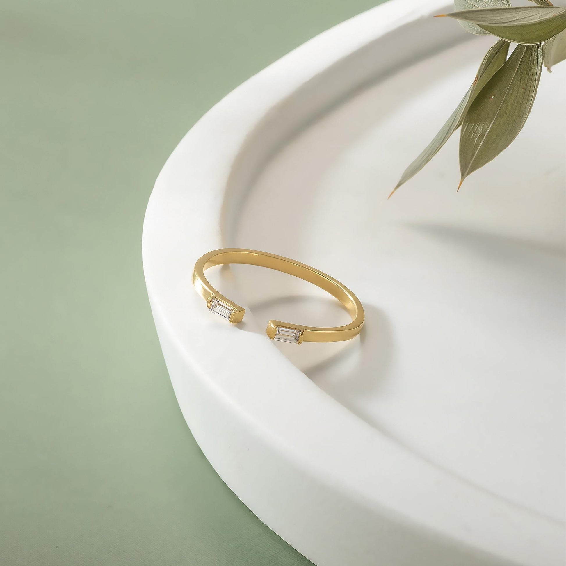 A gold ring with a unique design sits on a white surface, with a plant leaf visible in the background.