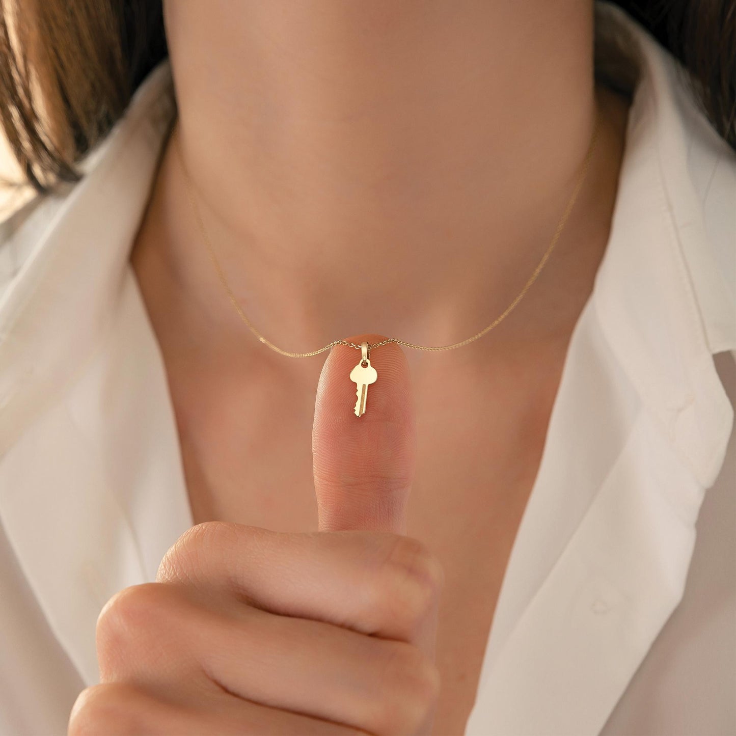 A person wearing a gold necklace with a small key charm on it, holding up their finger to show the charm.