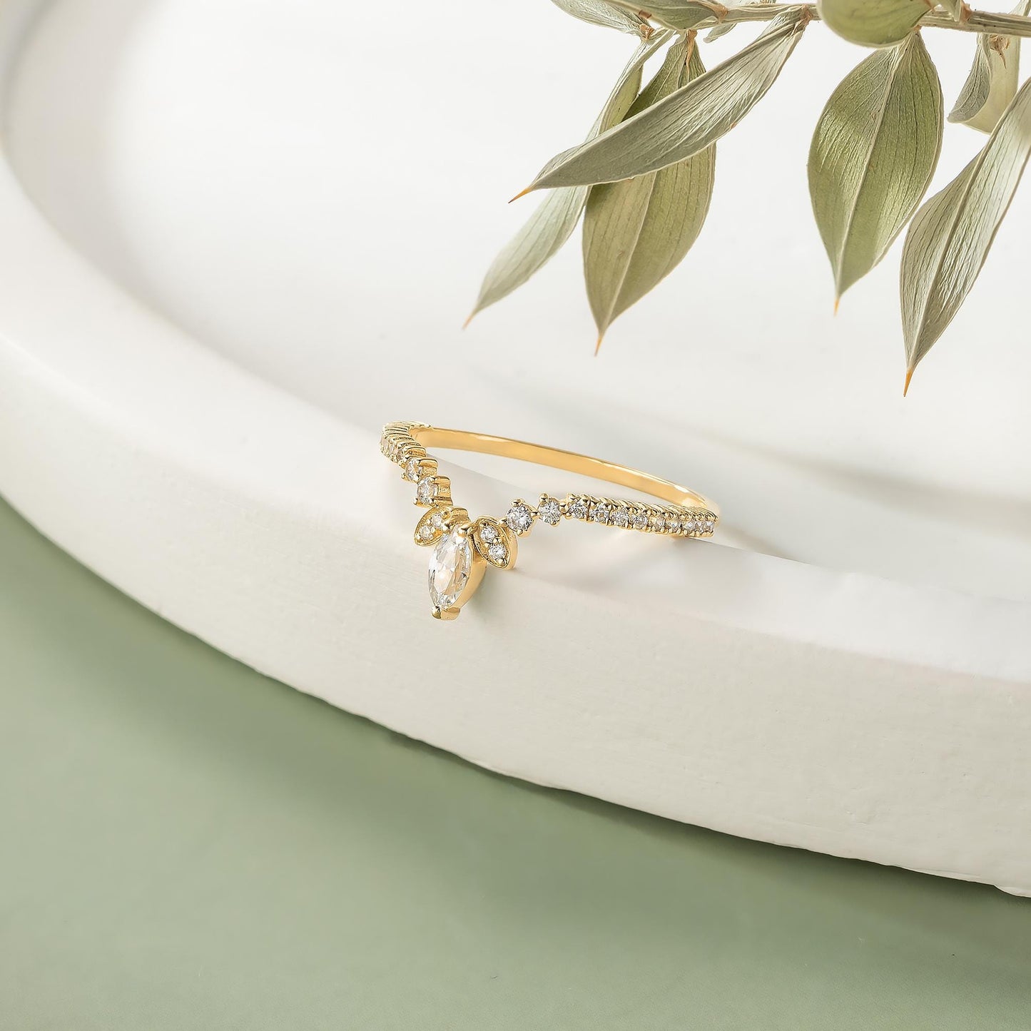 A gold ring with a large diamond in the center is displayed on a white surface, with a branch of green leaves in the background.