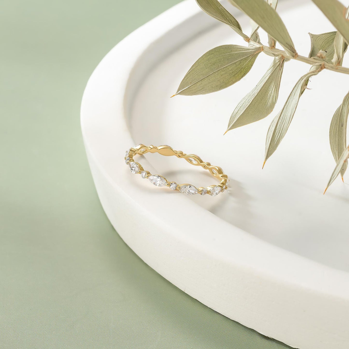A gold ring with a diamond-encrusted band is displayed on a white surface, with a branch of green leaves in the background.