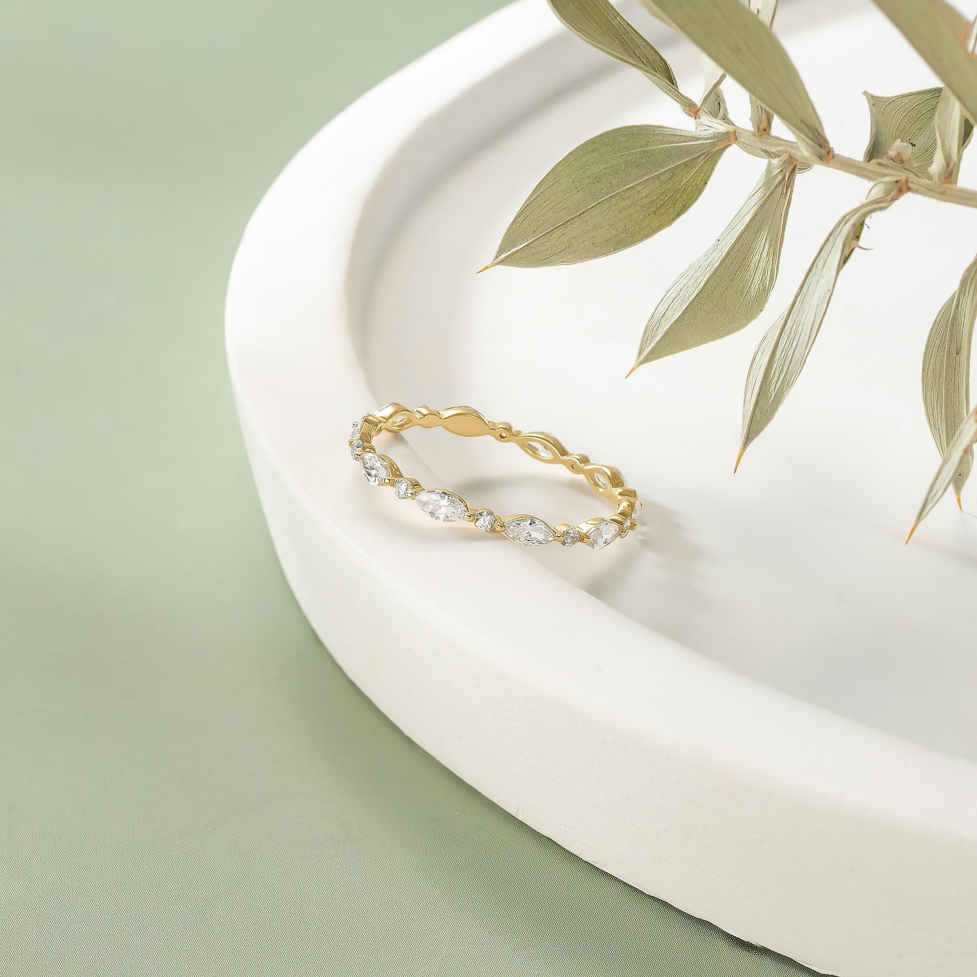 A gold ring with a diamond-encrusted band is displayed on a white surface, with a branch of green leaves in the background.