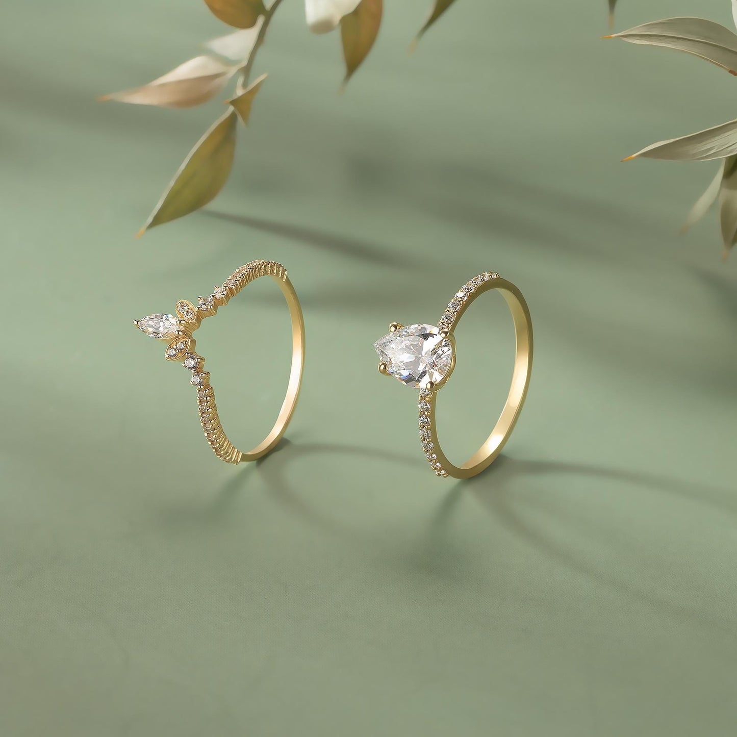 Two gold rings with diamond accents are displayed on a green background, with a branch of leaves in the background.