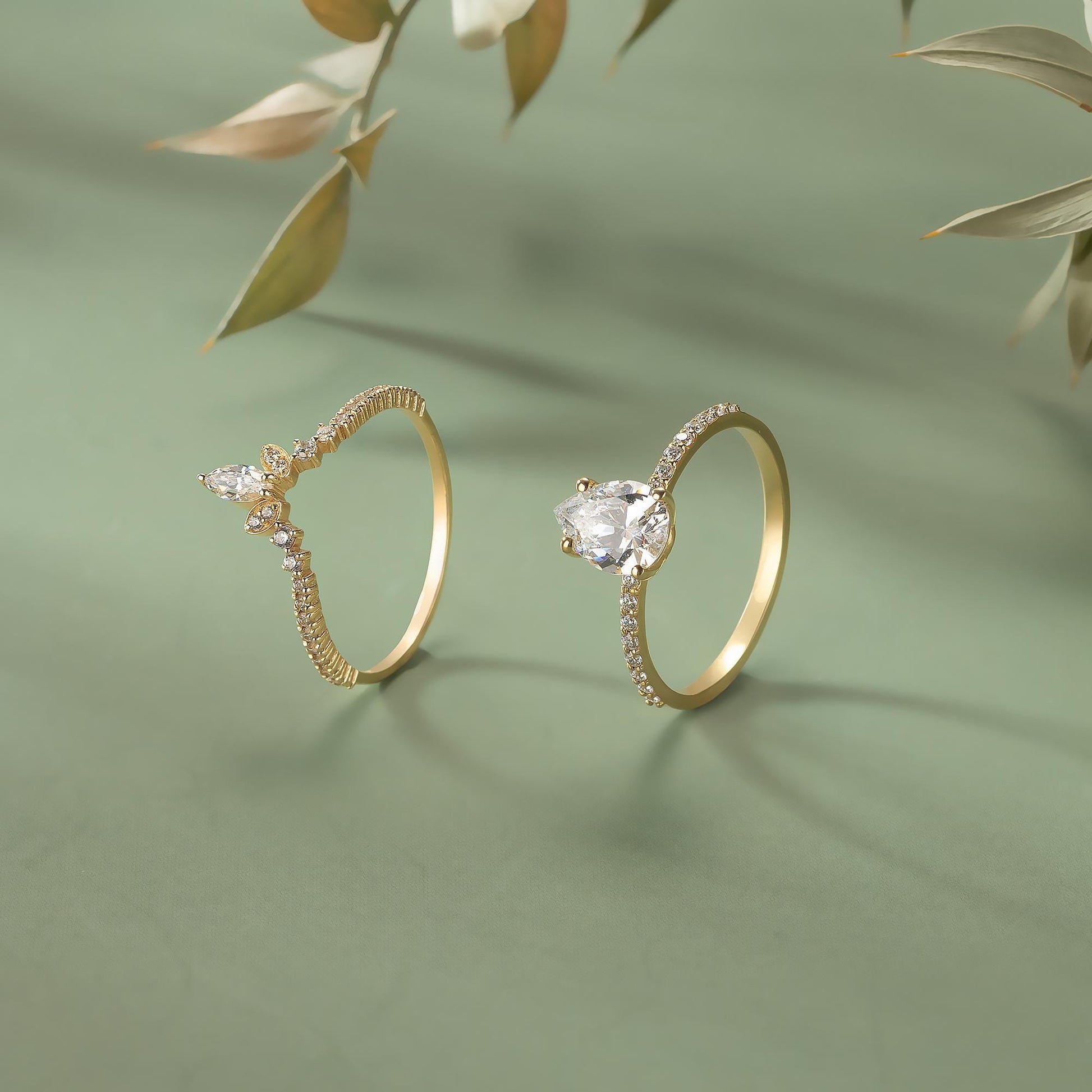 Two gold rings with diamond accents are displayed on a green background, with a branch of leaves in the background.