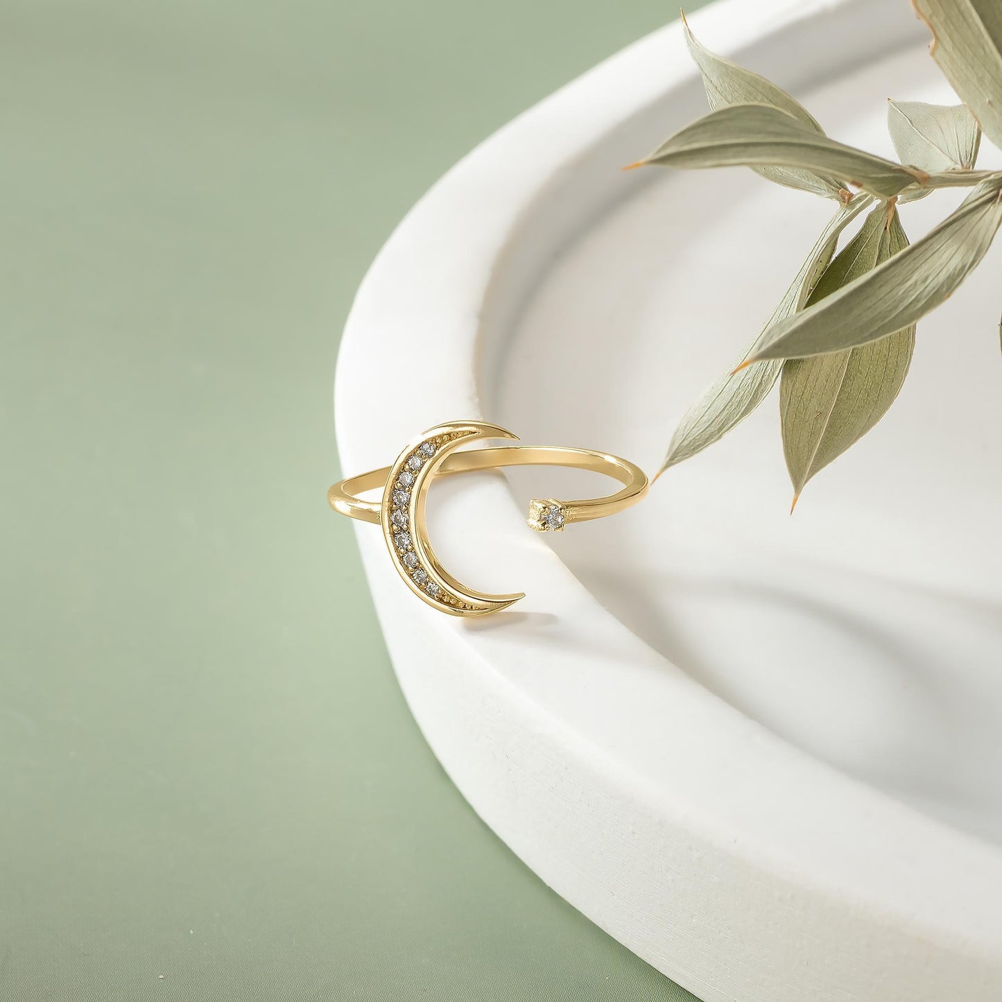 A gold ring with a crescent moon design and a small gemstone is displayed on a white surface, accompanied by a plant leaf.