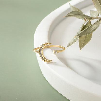 A gold ring with a crescent moon design and a small gemstone is displayed on a white surface, accompanied by a plant leaf.