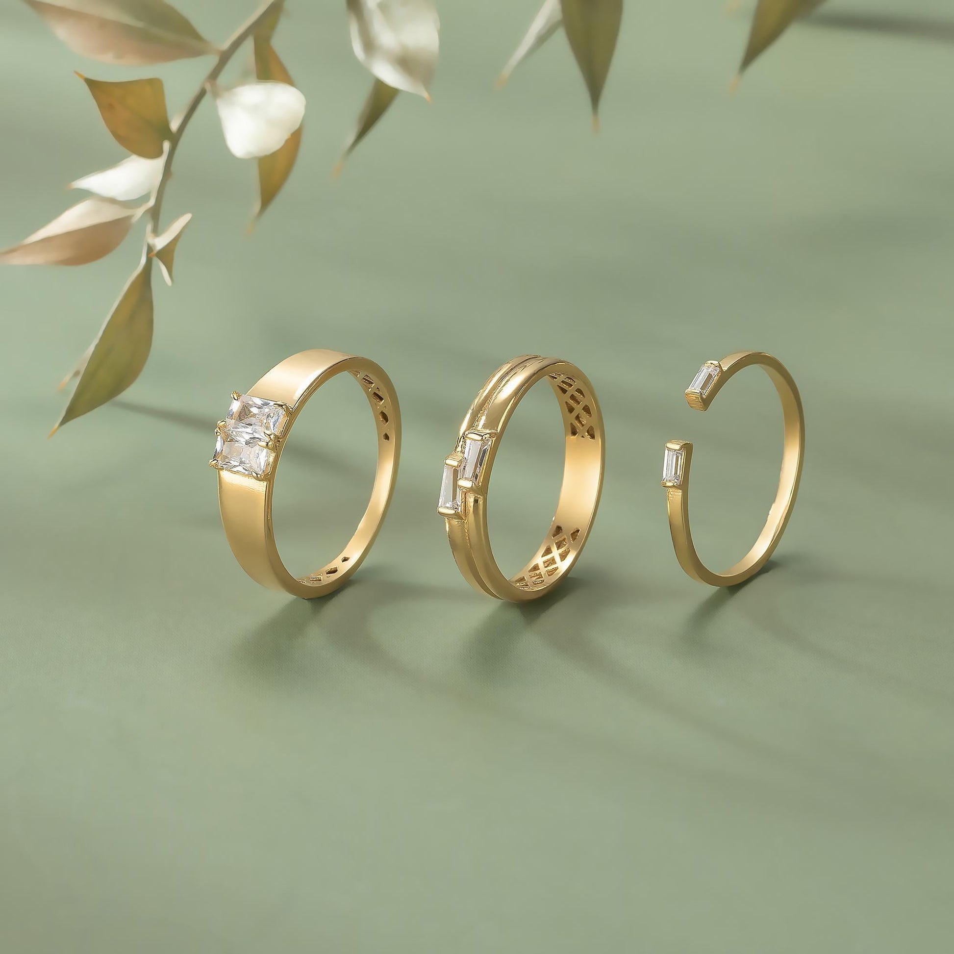 Three gold rings with gemstones are displayed against a green background, with a branch of leaves in the background.