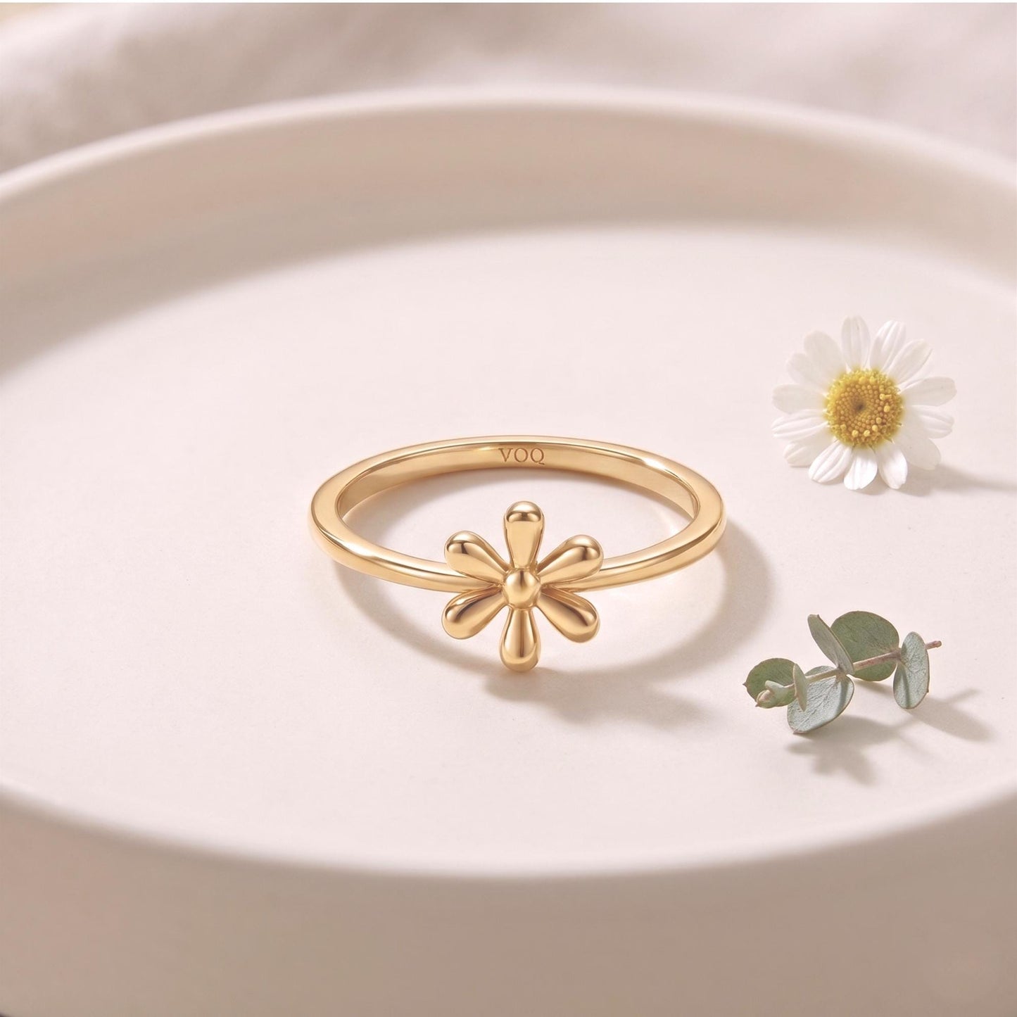 A gold ring with a flower design sits on a white surface, accompanied by a single white daisy flower and some greenery.