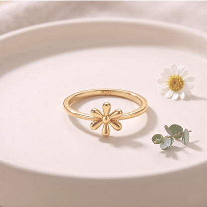 A gold ring with a flower design sits on a white surface, accompanied by a single white daisy flower and some greenery.