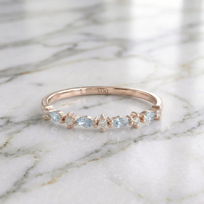 A rose gold ring with a row of aquamarine marquise and round moissanite stones stones set in a diamond pattern, resting on a marble surface.