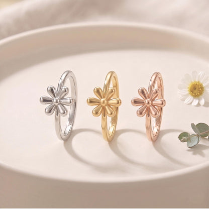 Three rings with flower designs are displayed on a white surface, accompanied by a single white flower and some greenery.