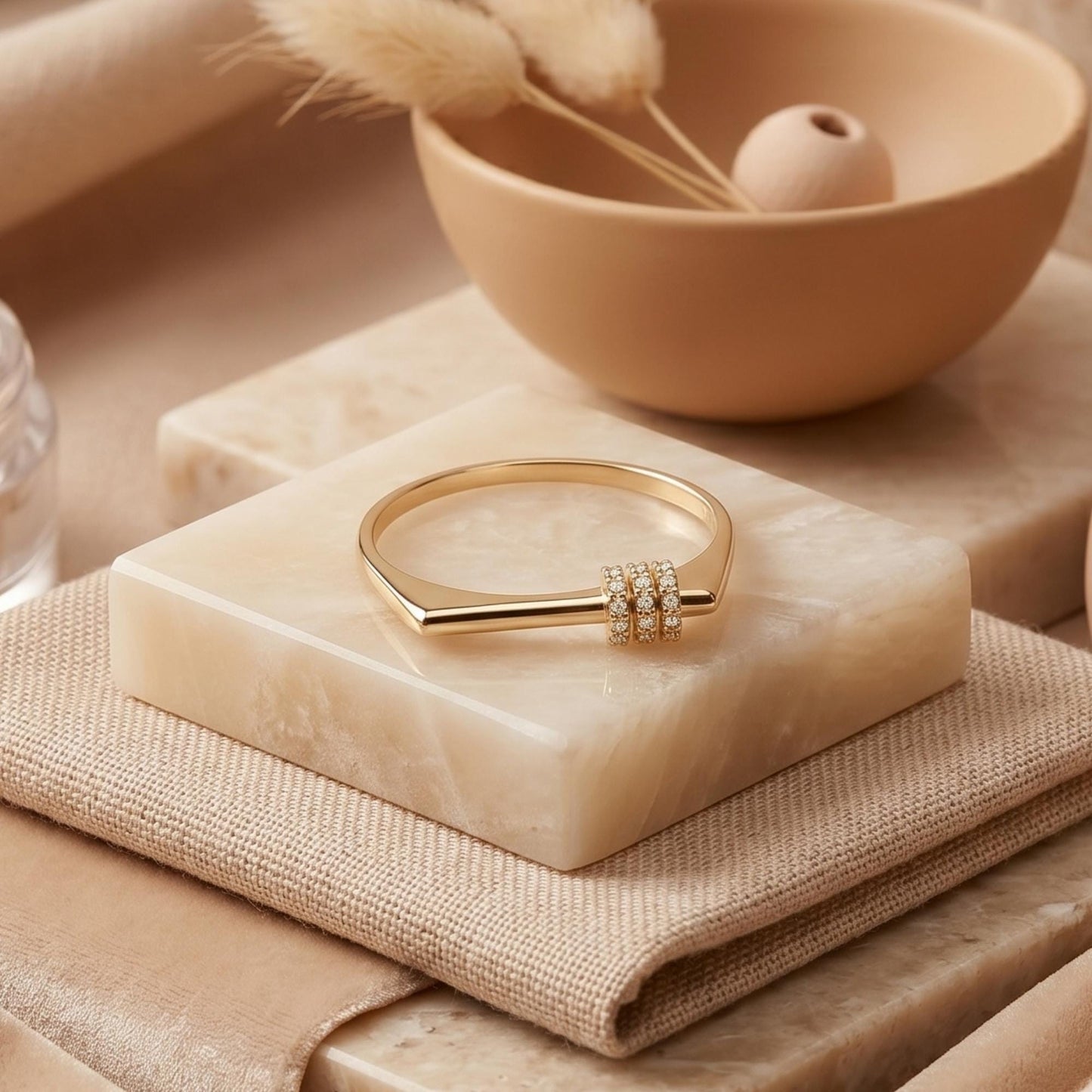 a gold ring with a diamond accent, placed on a marble surface, with a bowl of dried flowers and a beige bowl in the background.