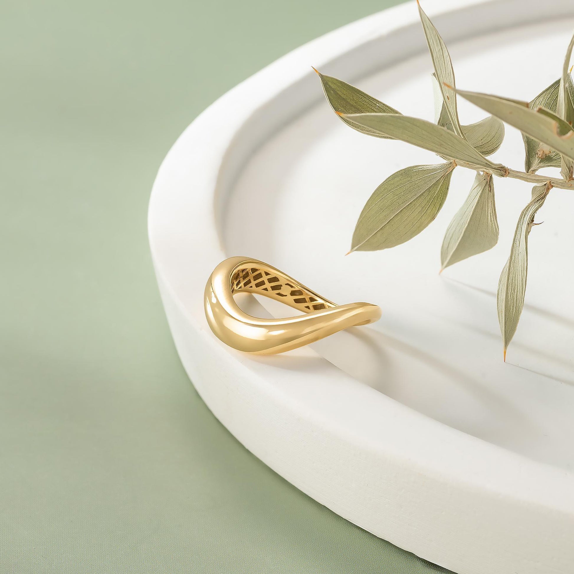 a gold ring with a unique design resting on a white plate, accompanied by a branch of green leaves.