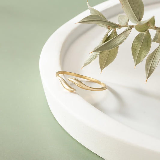 A gold ring with a leaf-like design rests on a white plate, accompanied by a branch of green leaves.