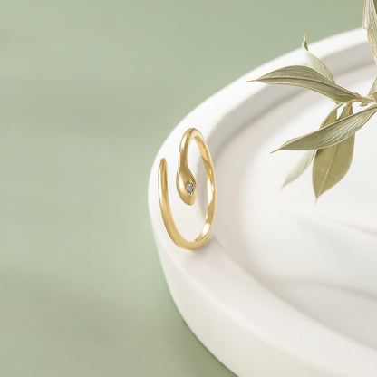 A gold snake-shaped ring with a small diamond is displayed on a white plate, accompanied by a leafy plant.