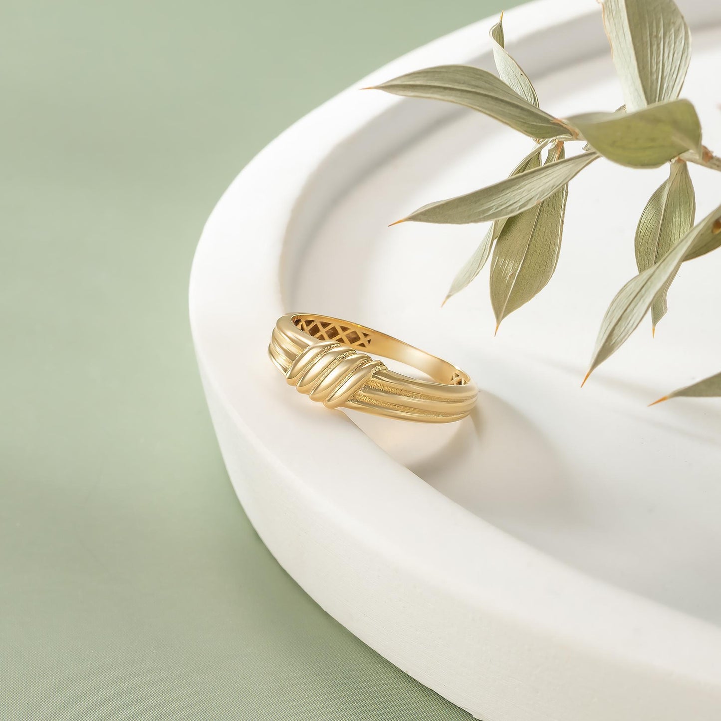 A gold ring with a twisted band sits on a white plate, accompanied by a branch of green leaves.