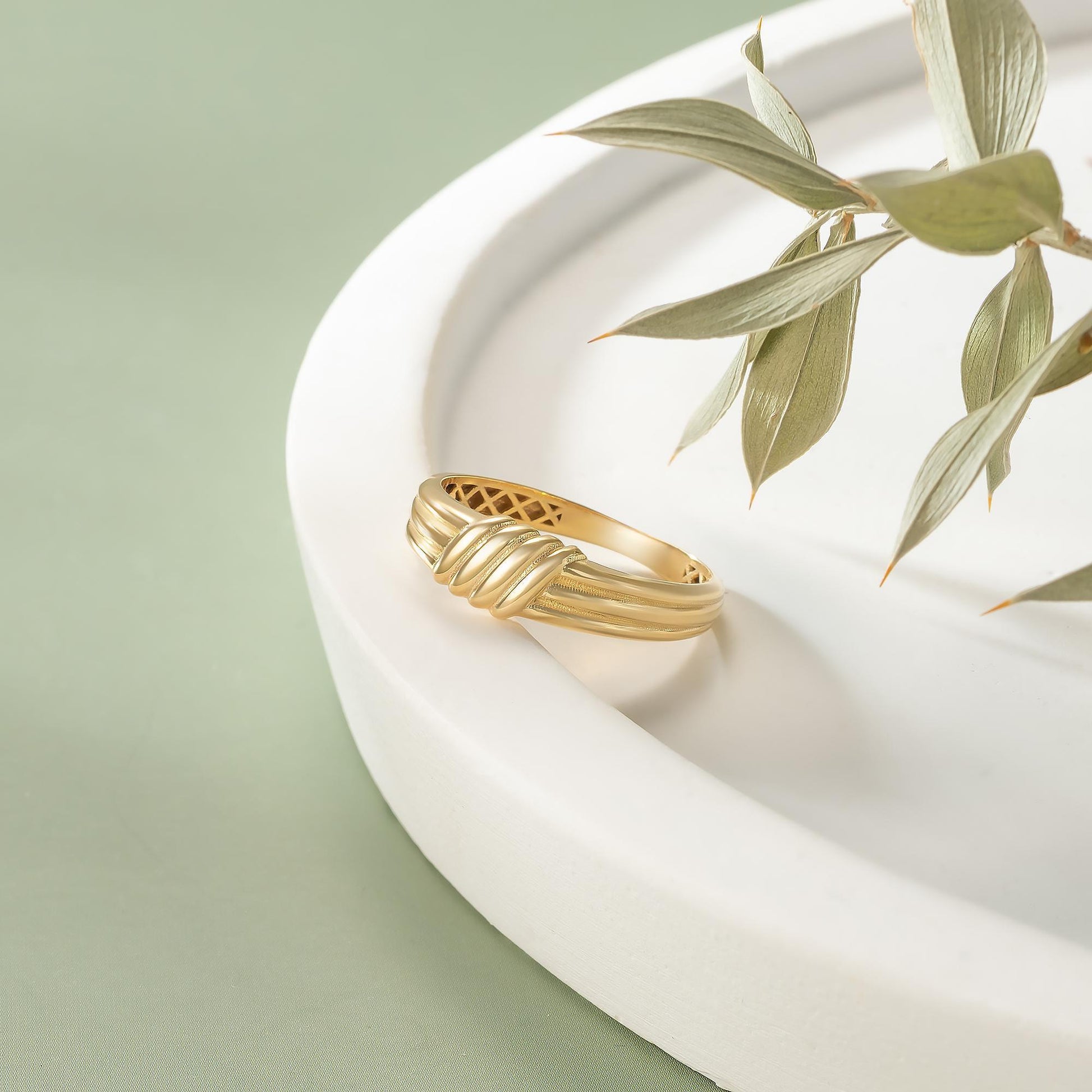 A gold ring with a twisted band sits on a white plate, accompanied by a branch of green leaves.