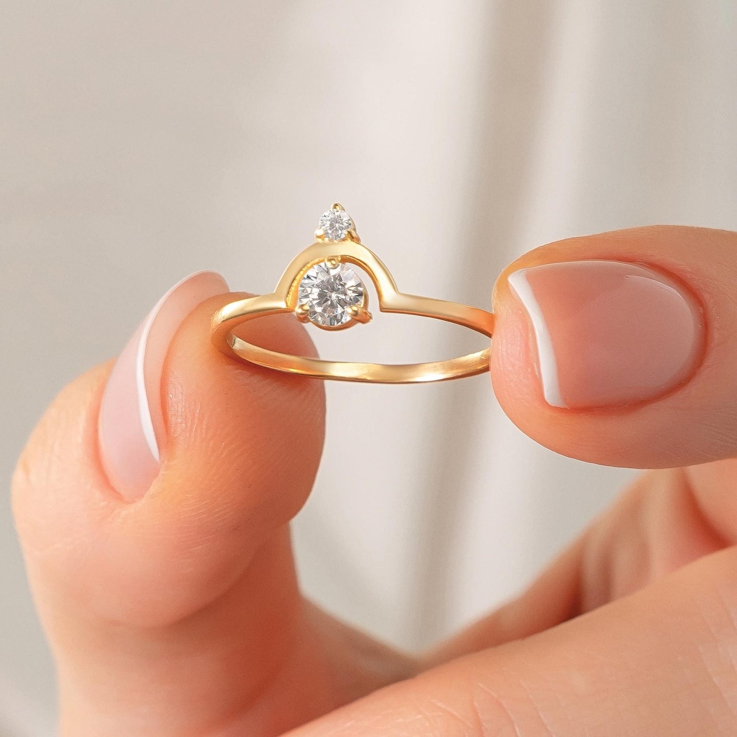 May include: A gold ring featuring a large, round, clear gemstone and a smaller gemstone above it. The ring has a unique arched design. The ring is held between two fingers.
