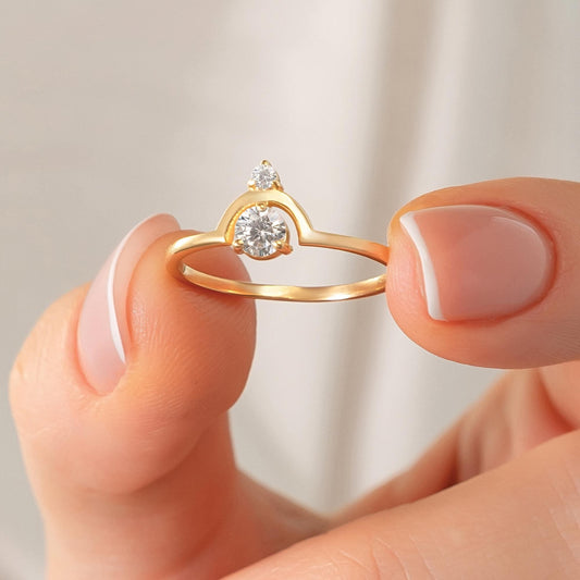 May include: A gold ring featuring a large, round, clear gemstone and a smaller gemstone above it. The ring has a unique arched design. The ring is held between two fingers.