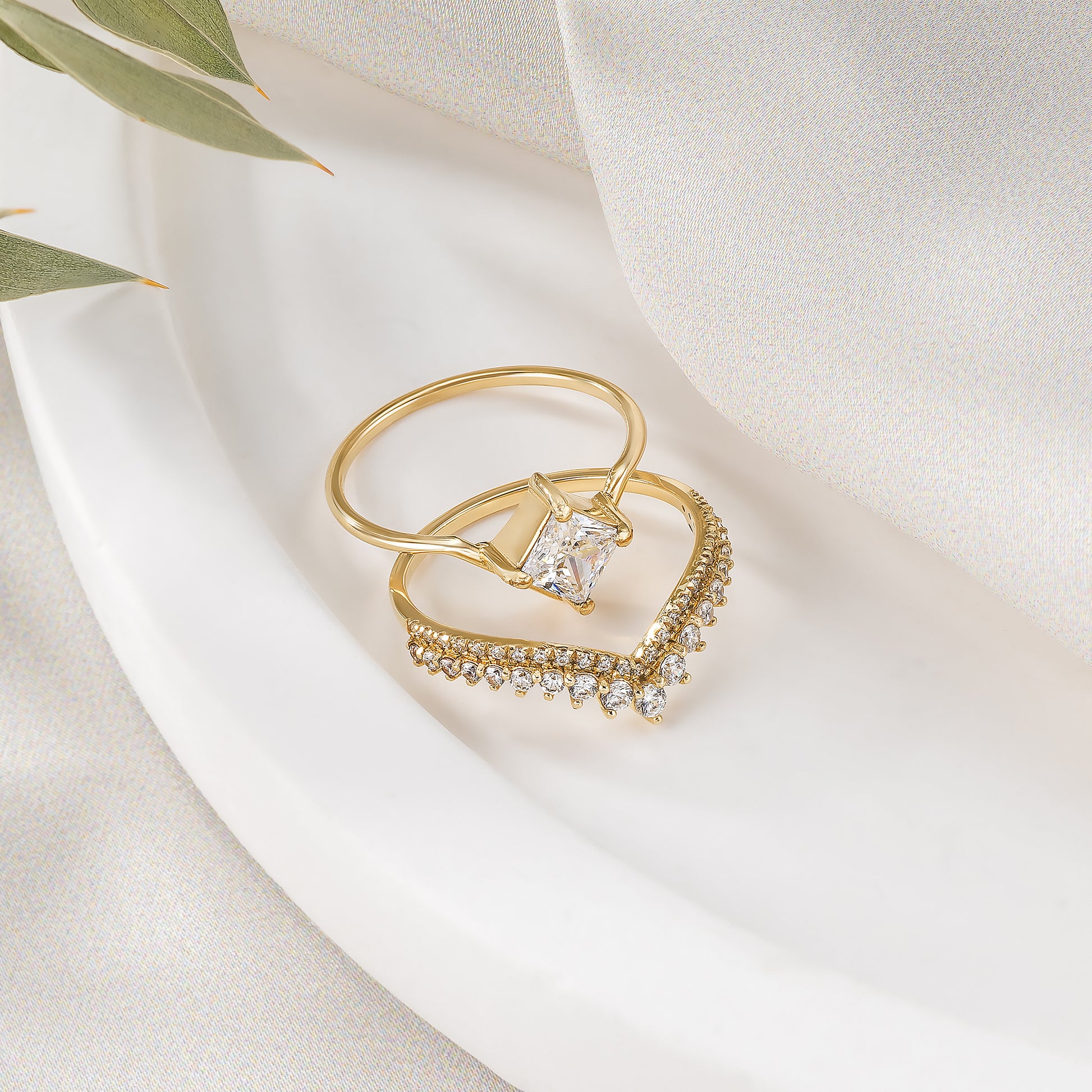 A gold ring with a diamond and a diamond-encrusted ring are displayed on a white cloth, with a green leaf visible in the background.