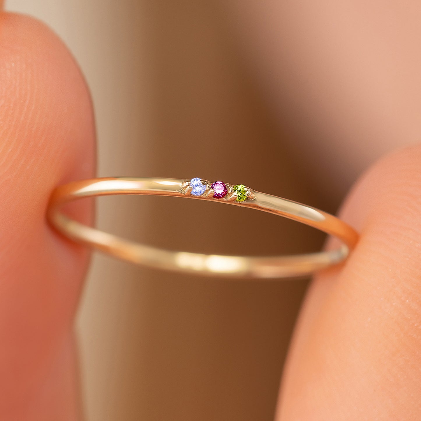 A close-up view of a gold ring with three gemstones, including a blue gemstone, a pink gemstone, and a green gemstone, being held between two fingers.
