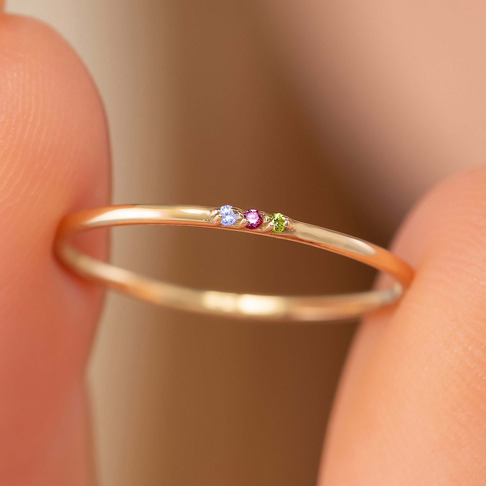 A close-up view of a gold ring with three gemstones, including a blue gemstone, a pink gemstone, and a green gemstone, being held between two fingers.