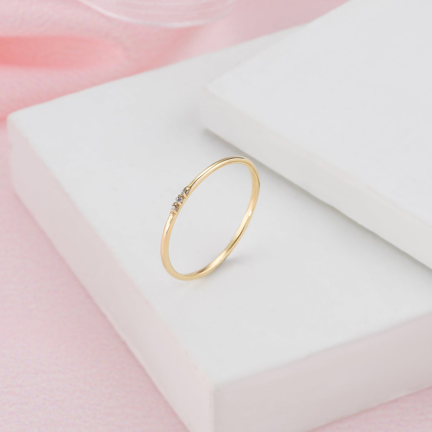 A delicate gold ring with a small gemstone is displayed on a white surface against a pink background.