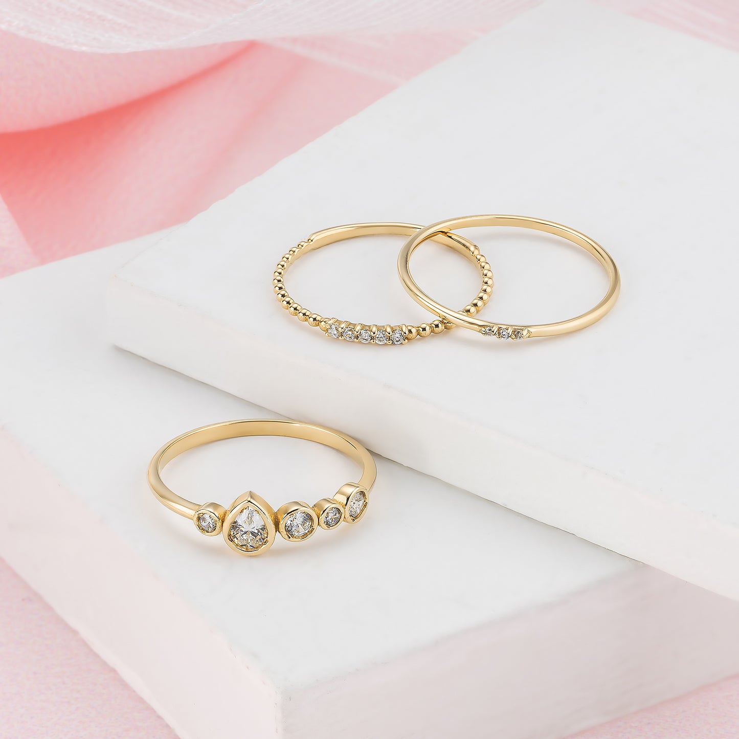 Three gold rings with diamonds are displayed on a white surface against a pink background.