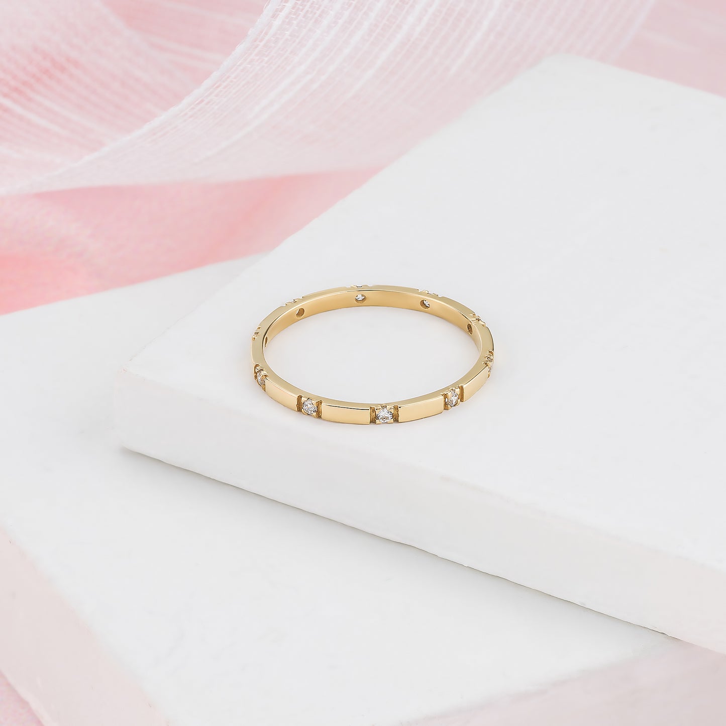 A gold ring with small diamonds is displayed on a white surface against a pink background.
