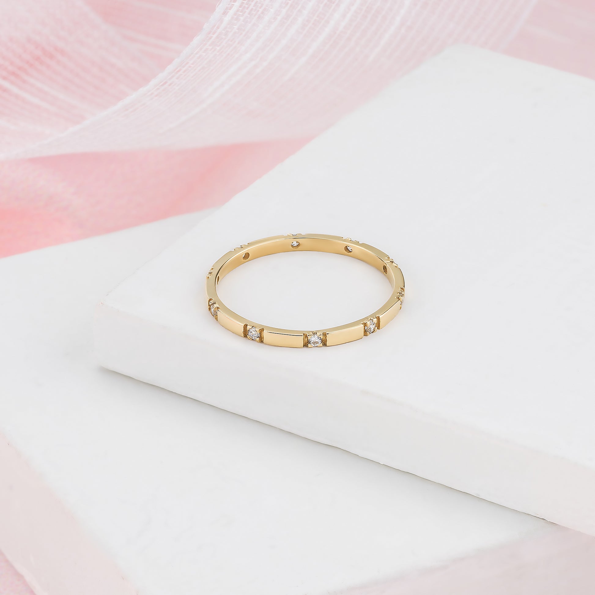 A gold ring with small diamonds is displayed on a white surface against a pink background.