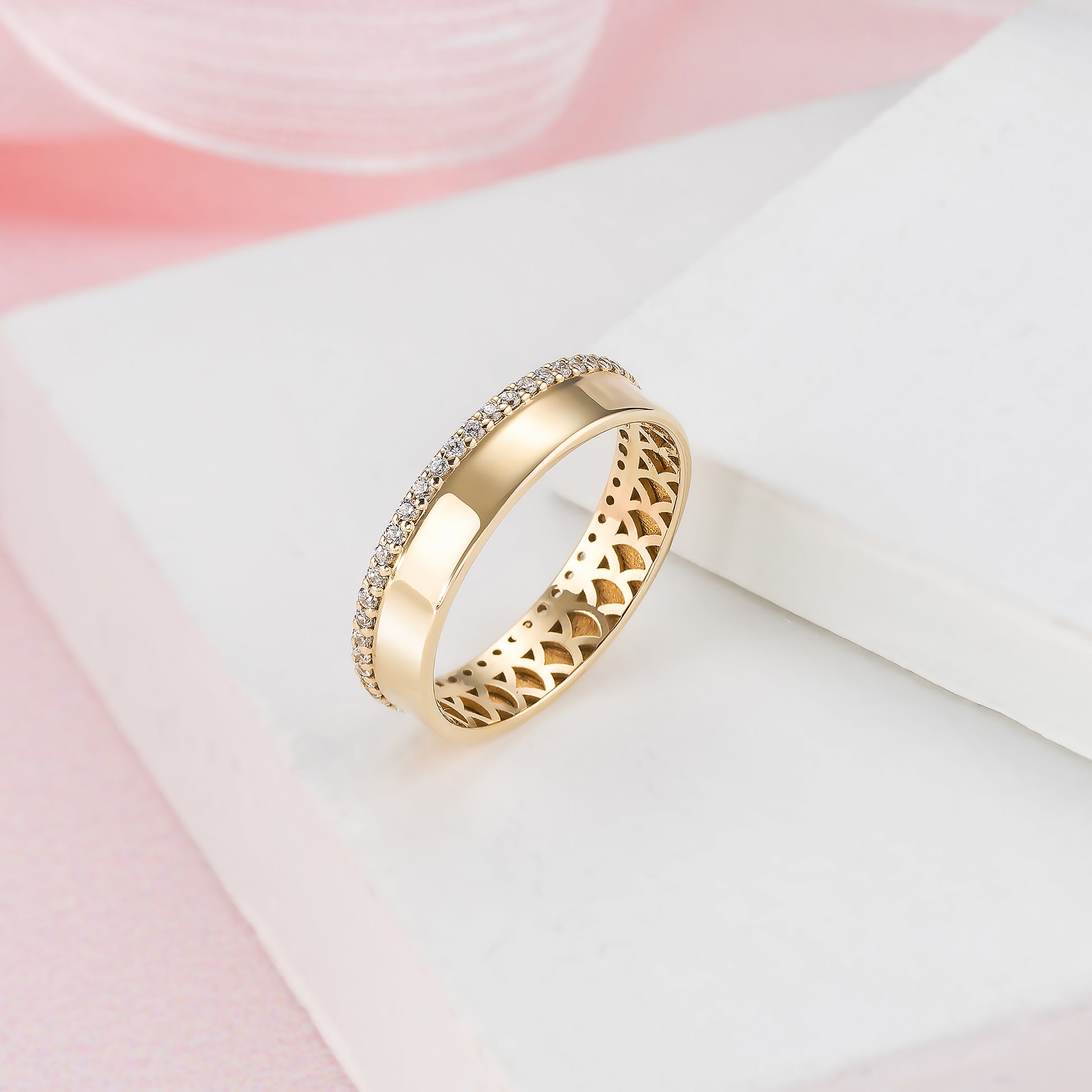 A gold ring with a diamond-encrusted band is displayed on a white surface against a pink background.