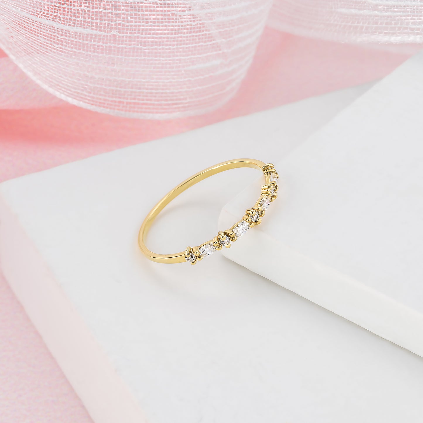 A gold ring with a decorative band of small diamonds is displayed on a white surface against a pink background.