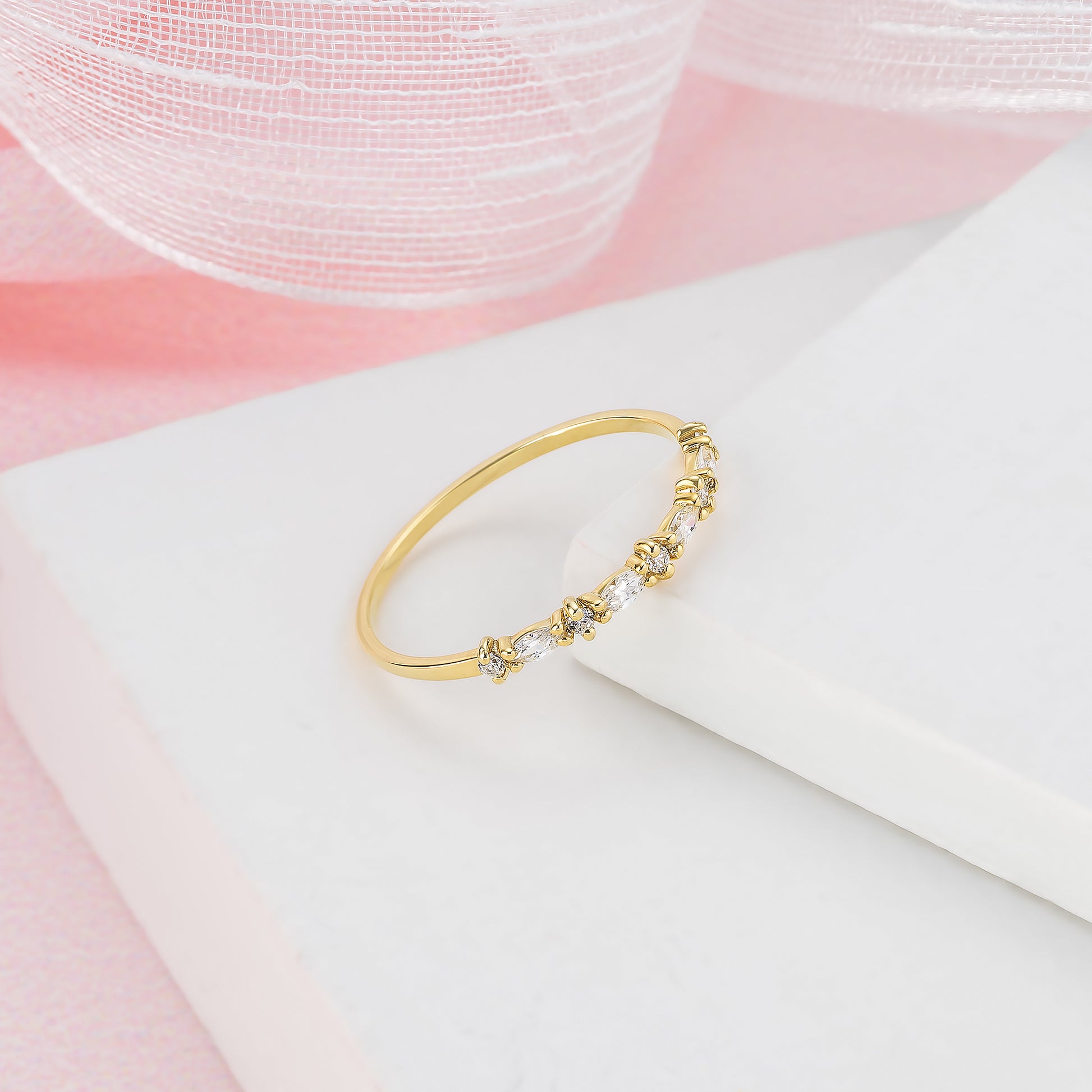 A gold ring with a decorative band of small diamonds is displayed on a white surface against a pink background.