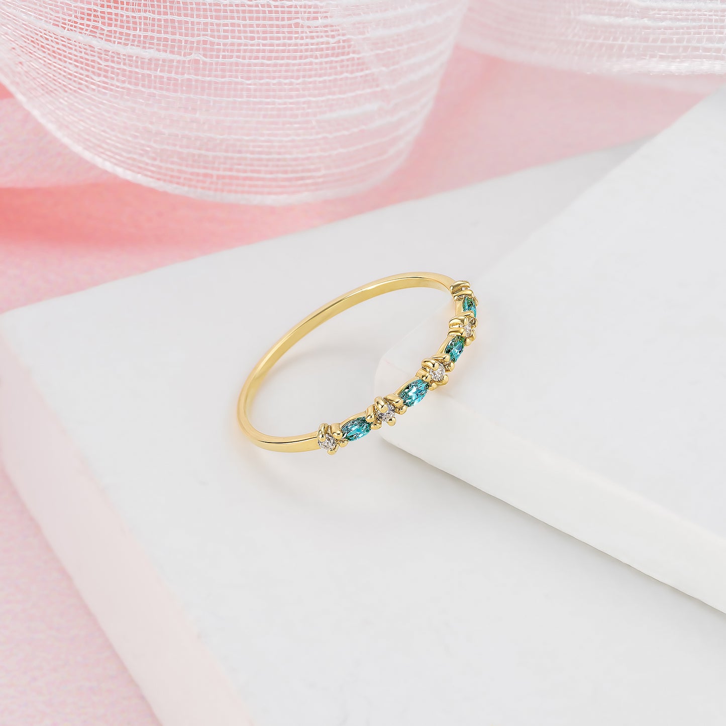 A gold ring with blue and green gemstones is displayed on a white surface against a pink background.