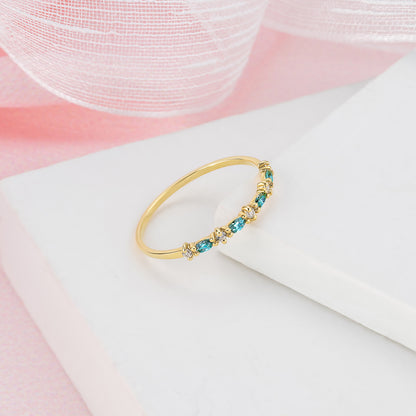 A gold ring with blue and green gemstones is displayed on a white surface against a pink background.