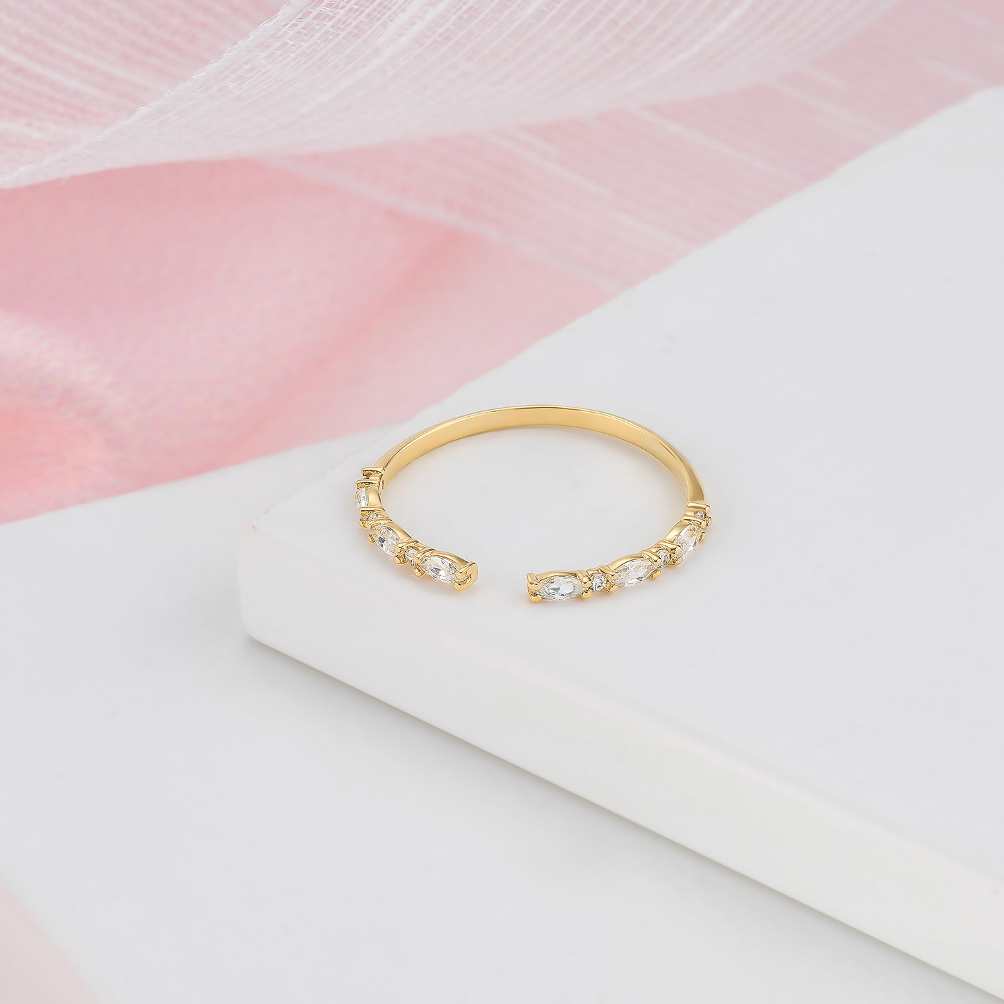 A gold ring with a row of small diamonds on the band is displayed on a white surface against a pink background.