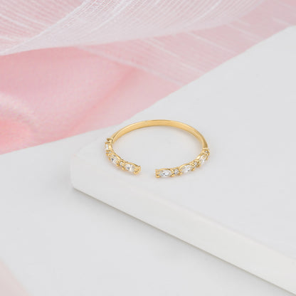 A gold ring with a row of small diamonds on the band is displayed on a white surface against a pink background.