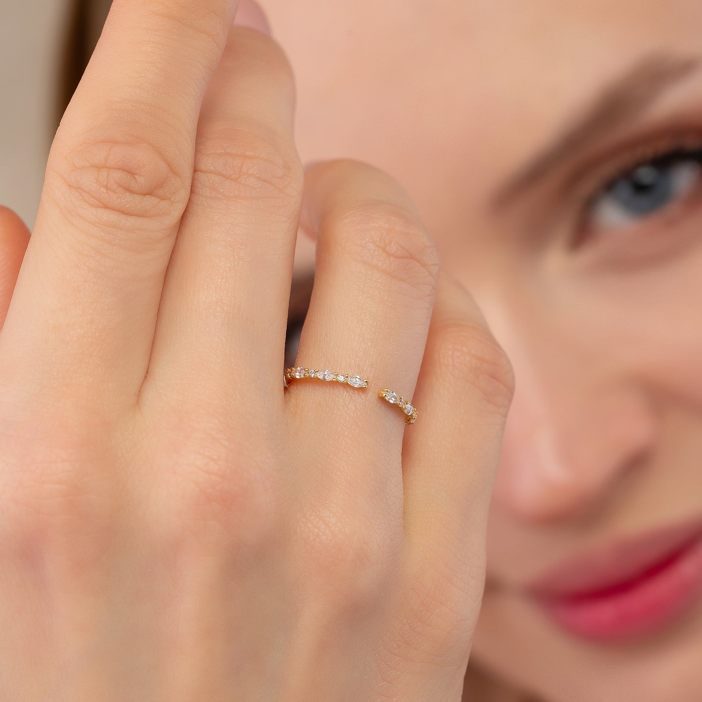 A close-up of a person's hand wearing a gold ring with small diamonds, with a blurred background of a person's face.