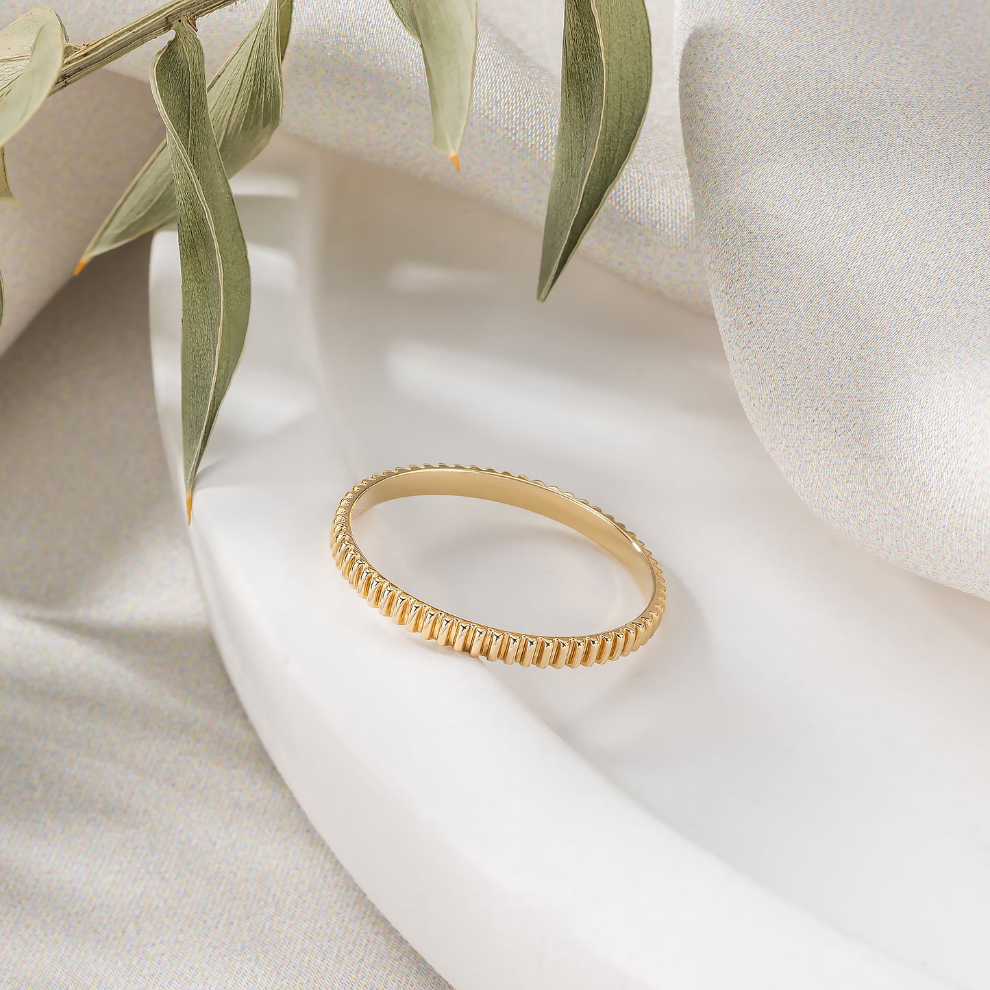 A gold ring with a textured band is displayed on a white surface, accompanied by a green leaf.