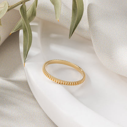 A gold ring with a textured band is displayed on a white surface, accompanied by a green leaf.