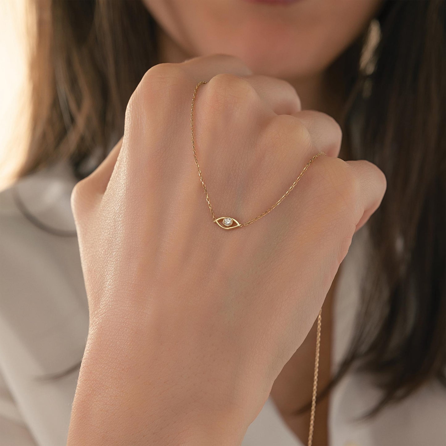 A close-up of a person's hand wearing a delicate gold necklace with a small eye-shaped pendant.