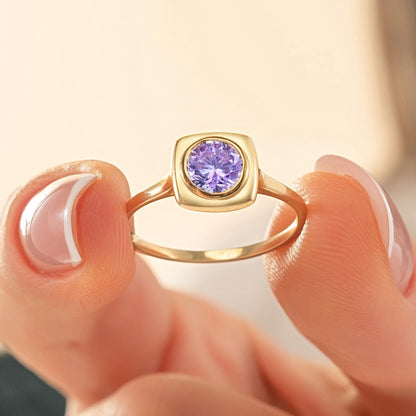 A close-up image of a person's hand holding a gold ring with a purple gemstone, specifically a purple sapphire.