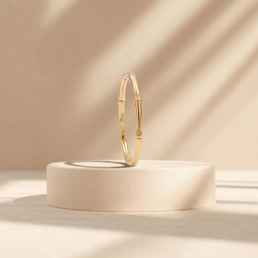 A gold bangle bracelet is displayed on a white pedestal against a light beige background.