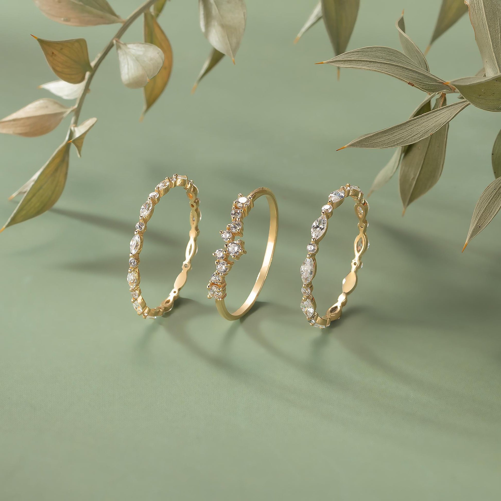 Three gold rings with intricate designs and sparkling gemstones are displayed against a green background, with some leaves visible in the background.
