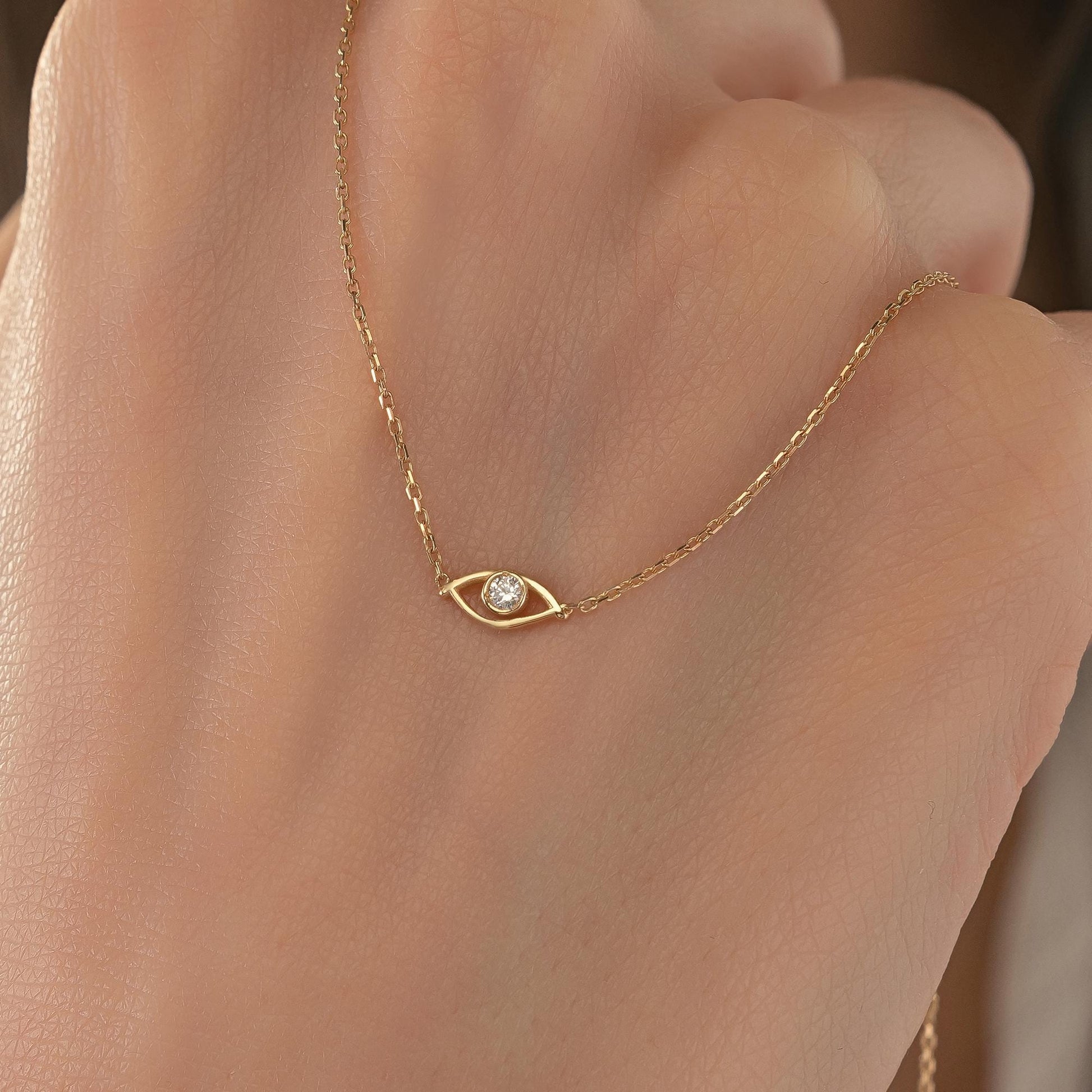 A close-up of a person's hand wearing a delicate gold necklace with a small diamond-encrusted eye pendant.