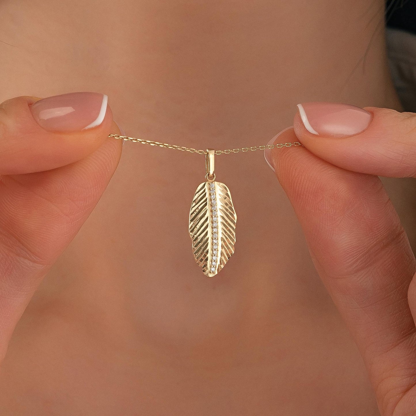 A person's hand holding a delicate gold feather pendant necklace.